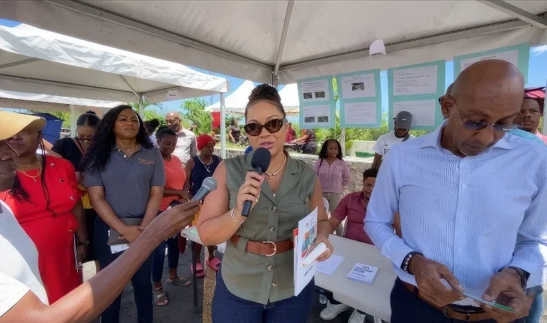 Melissa Skerrit speaks to the press with a microphone at a housing fair booth, surrounded by attendees and informational displays under a white tent.