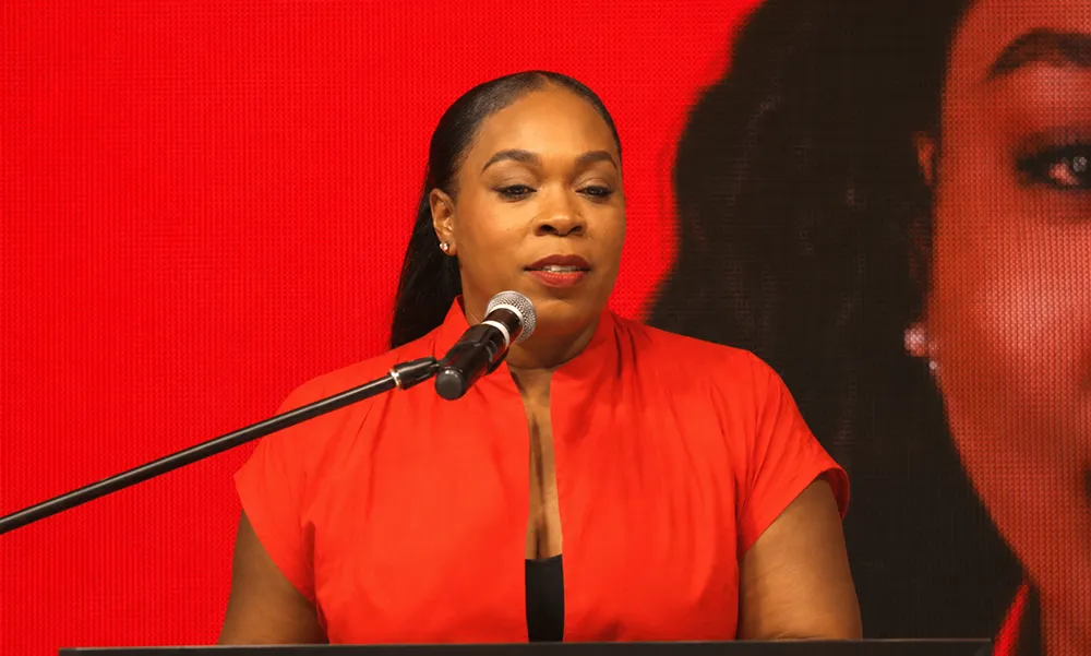 Close-up campaign image of Ashma McDougall, the DLP Roseau North political candidate, speaking at a podium in Lindo Park against a red stage backdrop with part of her enlarged portrait behind her.