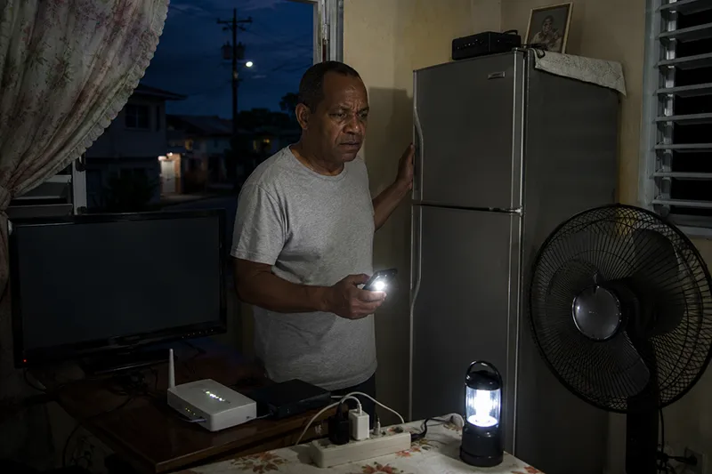 A Dominican homeowner stands in a dimly lit home checking appliances after repeated electricity outages, reflecting frustration over unstable power and appliance damage concerns.