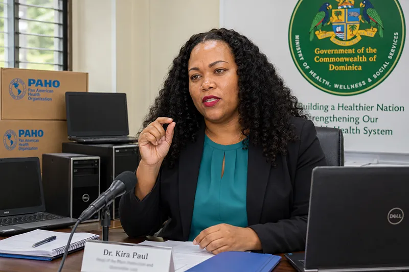 Dr. Kyra Paul speaks in a professional health office in Dominica, gesturing with her index finger and thumb while laptops, desktop units, PAHO-marked boxes, and the ministry logo are visible.