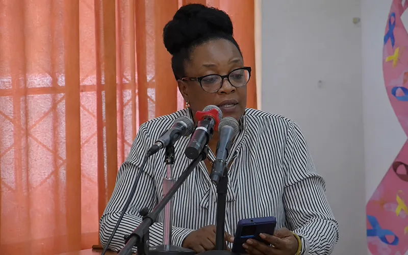 Dr. Velda Wade speaking at the Dominica Cancer Society Annual General Meeting in the same striped blouse and event setting seen in the reference images.