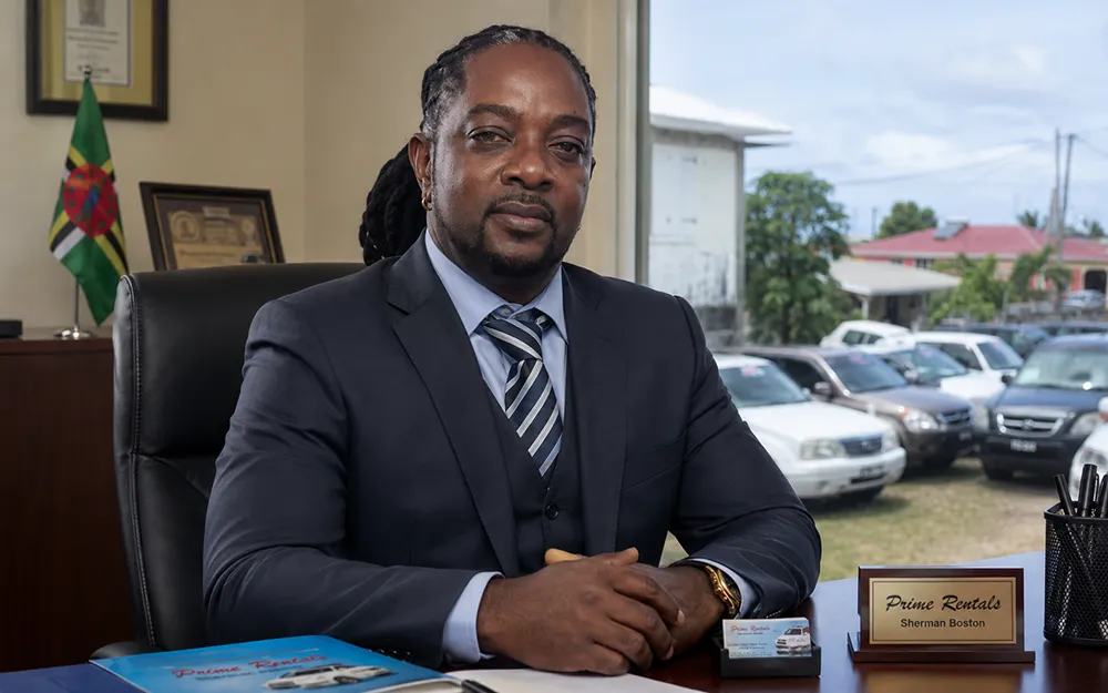 Sherman Boston seated formally in his office in Dominica with Prime Rentals branding and his rental vehicles visible through the window.