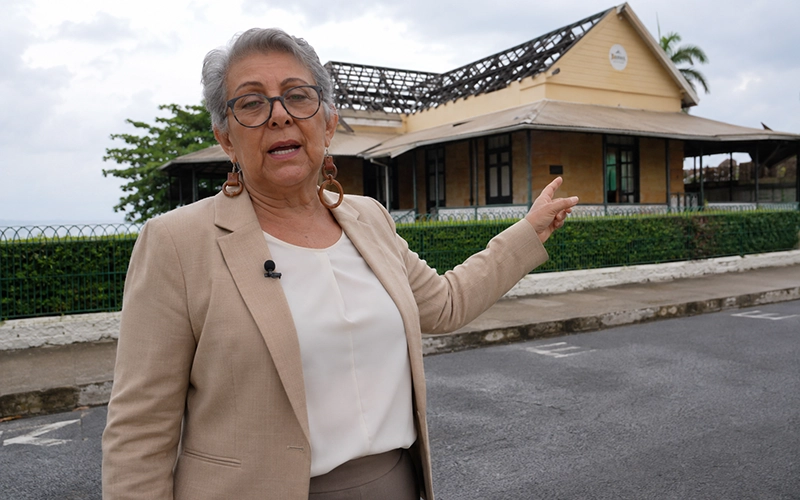 Anita Bully speaking in front of a damaged library building in Dominica while pointing at its deteriorating roof during an interview.