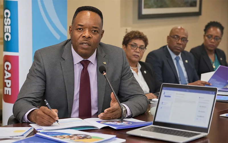CXC Registrar Dr. Wayne Wesley discusses School-Based Assessment reforms during a regional education briefing with assessment documents and a laptop before him.