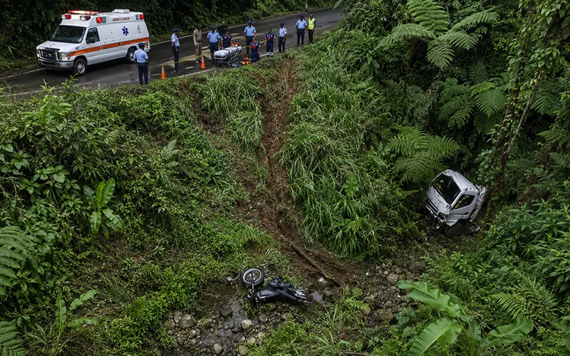 An aerial view shows police and an ambulance above a forested ravine where a damaged truck and overturned motorcycle came to rest.