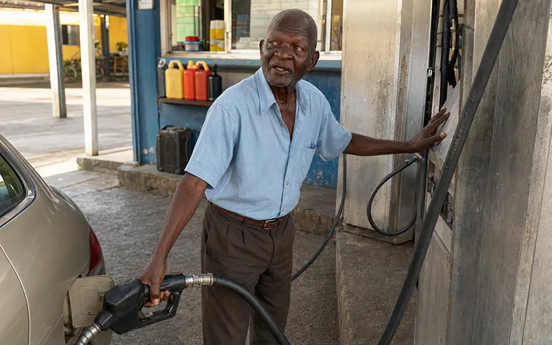 Veteran Dominican businessman McMillan “Mr. Mac” Jno-Baptiste holds a fuel nozzle while serving a vehicle at his modest roadside gas station.