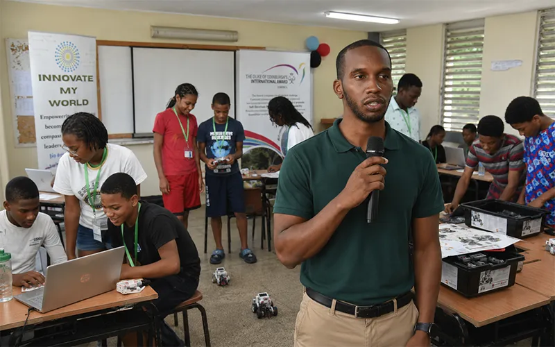 A robotics camp coordinator speaks in the foreground while Dominican secondary school students build, code and test educational robots inside a Portsmouth Secondary School classroom.
