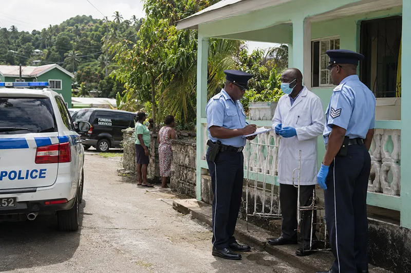 Police officers and a medical responder outside a modest home in Portsmouth, Dominica, during a respectful death investigation.