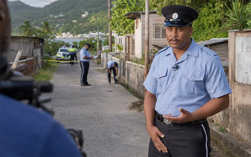 Police Public Relations Officer, dressed in a light blue Dominica police uniform and peaked cap with a clip-on microphone, gives an interview near a secured investigation area in Salisbury.